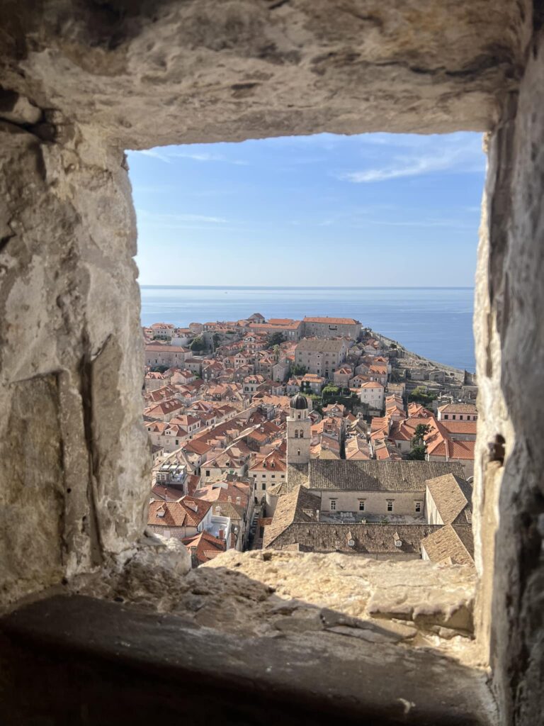 A view through a stone window shows a picturesque coastal town with terracotta rooftops and historic buildings, leading to a calm blue sea under a clear sky.