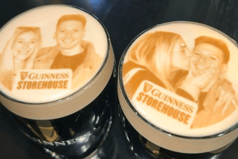 Two pints of Guinness with printed images on their foam. Each pint shows a sepia-toned photo of a smiling couple. The image on the left is a portrait, and the one on the right shows the woman playfully kissing the man on the cheek.