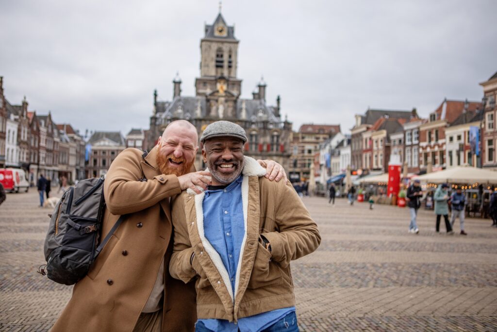 Two men pose happily in a cobblestone plaza with historic buildings in the background. One man playfully has his arm around the other's neck. Both are dressed in casual jackets and smiling broadly. It's a cloudy day.