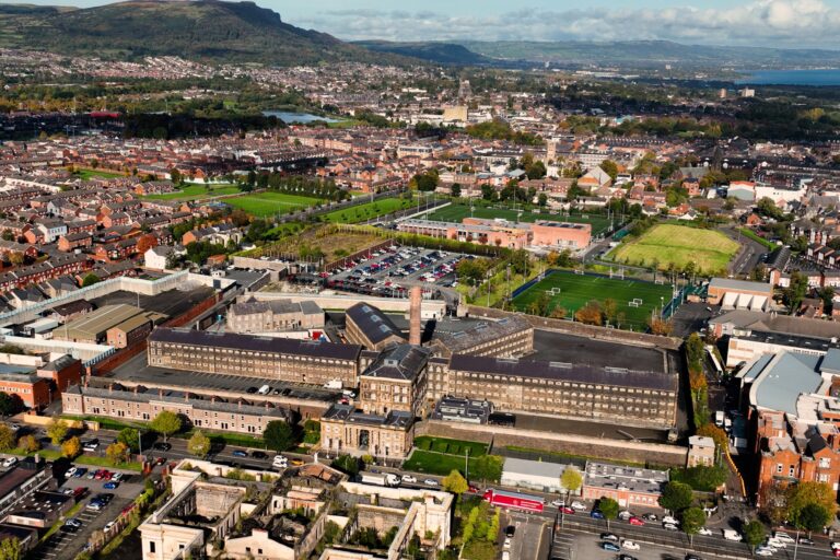 Aerial view of a large historic prison complex surrounded by urban landscape. The prison has distinctive stone buildings and a central courtyard. In the distance, residential neighborhoods and green hills are visible under a partly cloudy sky.