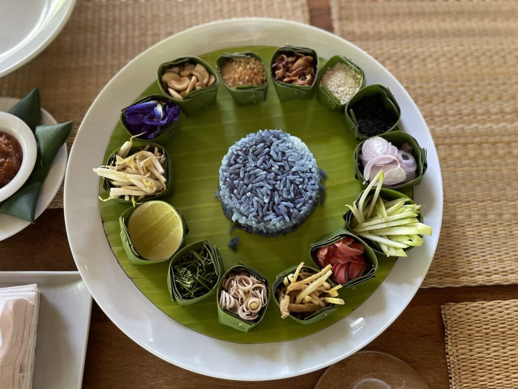 A beautifully arranged Thai dish featuring butterfly pea rice in the center, surrounded by various colorful ingredients in banana leaf cups, including sliced vegetables, herbs, and nuts, on a green banana leaf-covered plate.