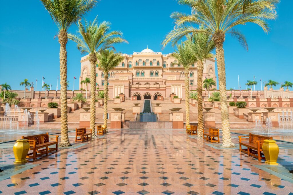 Grand, luxurious palace with a sandy-colored exterior, surrounded by palm trees and fountains. Polished tiled walkway with benches and large decorative urns leads to the entrance. Bright blue sky in the background.