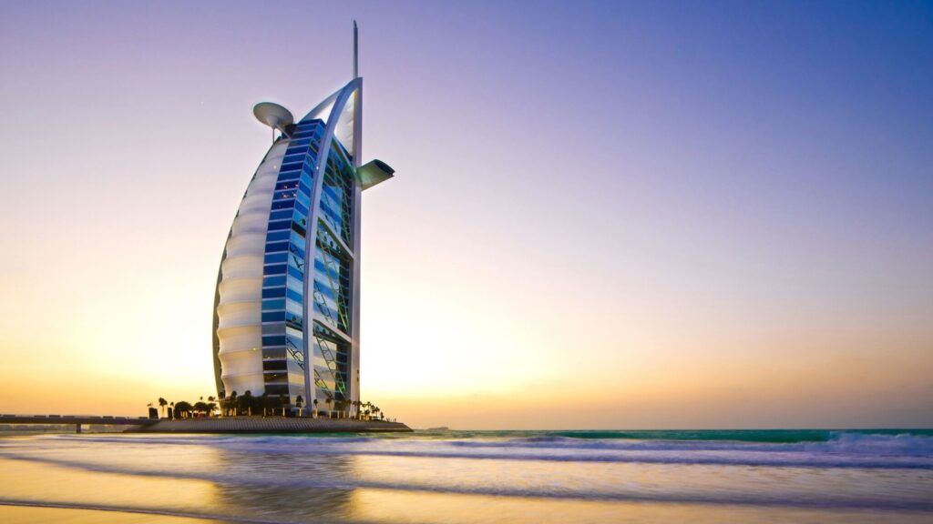 The image shows the Burj Al Arab, a luxury hotel in Dubai, shaped like a sail. It stands on an artificial island against a backdrop of a clear sky and sunset. The waves of the sea gently touch the shore in the foreground.