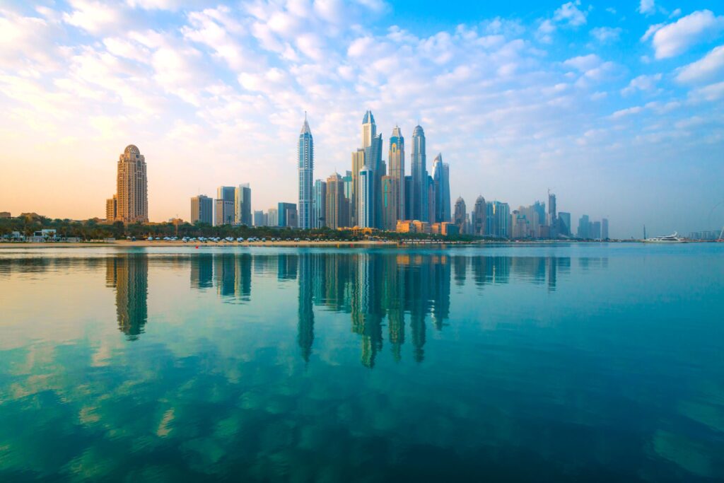 A city skyline with numerous skyscrapers reflects on calm water under a bright blue sky with scattered clouds. The sun casts a warm glow on the buildings, highlighting their modern architecture.