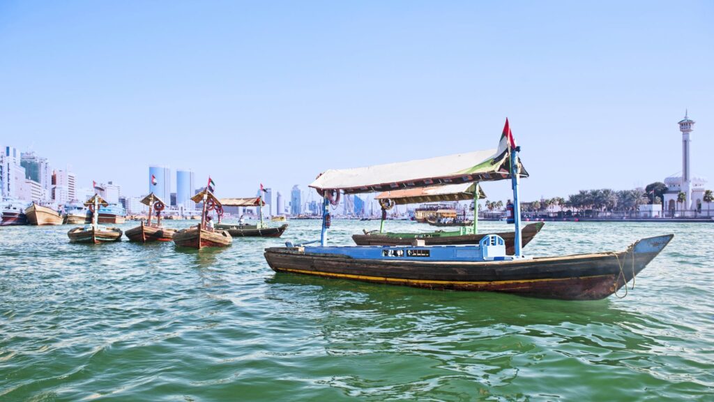 Traditional wooden boats with canopies float on a calm body of water, with a city skyline in the background. Clear blue sky above, a mosque with a tall minaret visible on the right side.