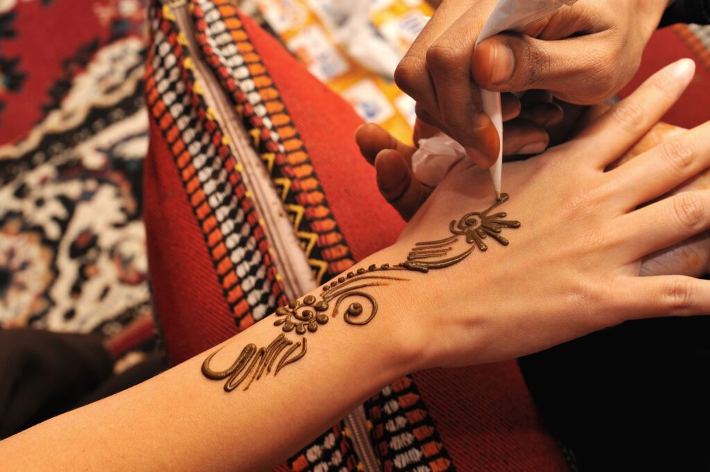 A close-up of a person applying intricate henna designs on someone's hand. The design features floral and geometric patterns, with the person's arm resting on a colorful fabric background.