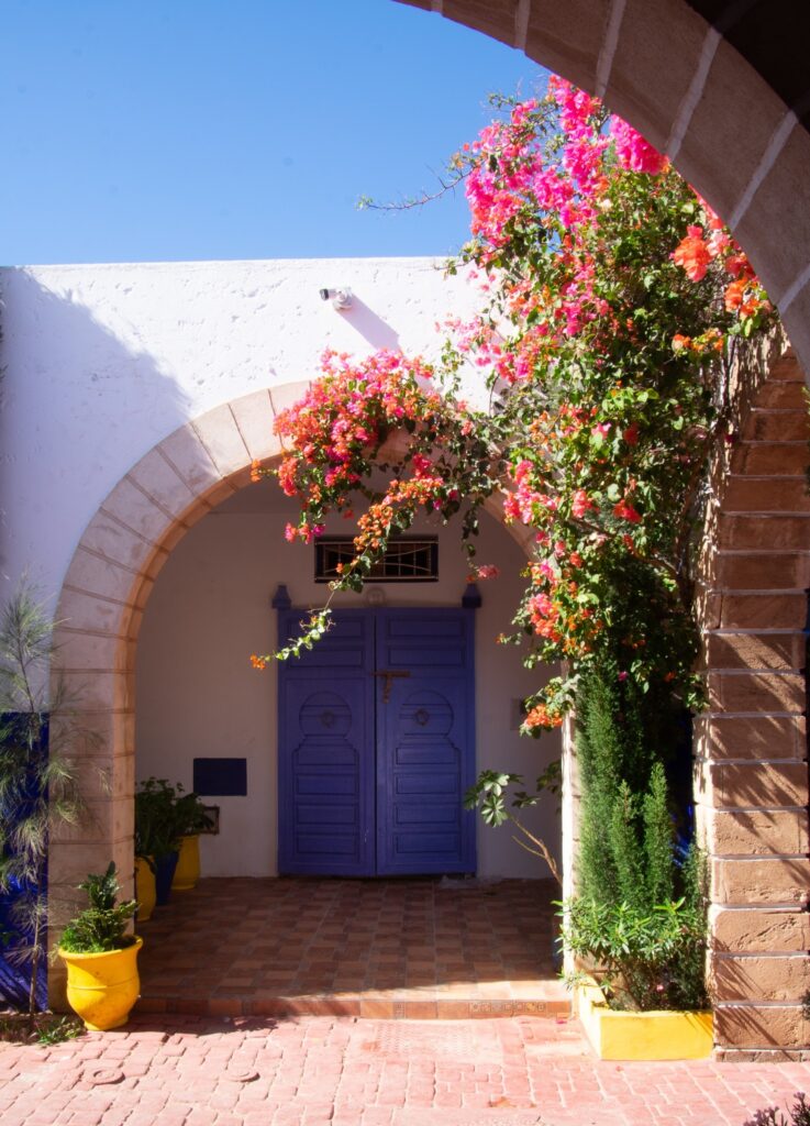 A vibrant scene of a building entrance with a vivid blue door framed by an arched stone doorway. Colorful bougainvillea climbs the wall, and yellow pots with green plants add contrast. The sky is clear and blue, adding to the serene atmosphere.